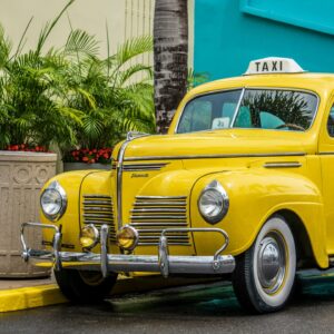 Vintage yellow taxi car parked on an urban street with lush green plants and palm trees.