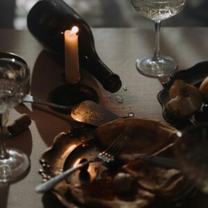 Moody candlelit table with wine bottle, empty plates, and scattered tableware, evoking a finished meal.